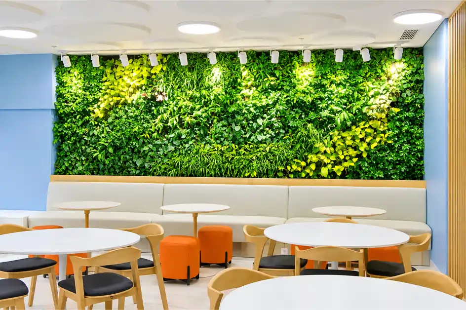 Indoor greenwall lebar dengan lampu sorot di atas sofa dan meja bundar area makan kafetaria